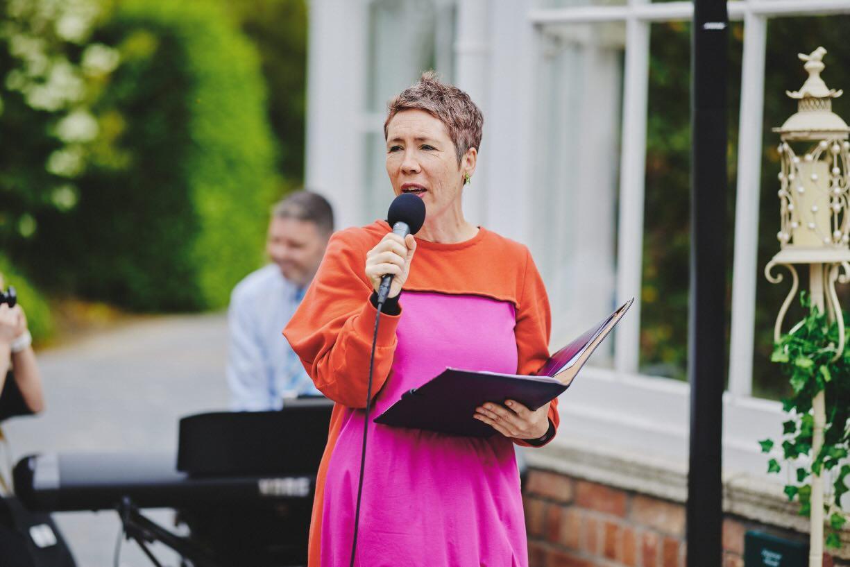 elebrant in a bright orange and pink dress speaking into a microphone and reading from a folder at an outdoor ceremony, with a keyboard player in the background
